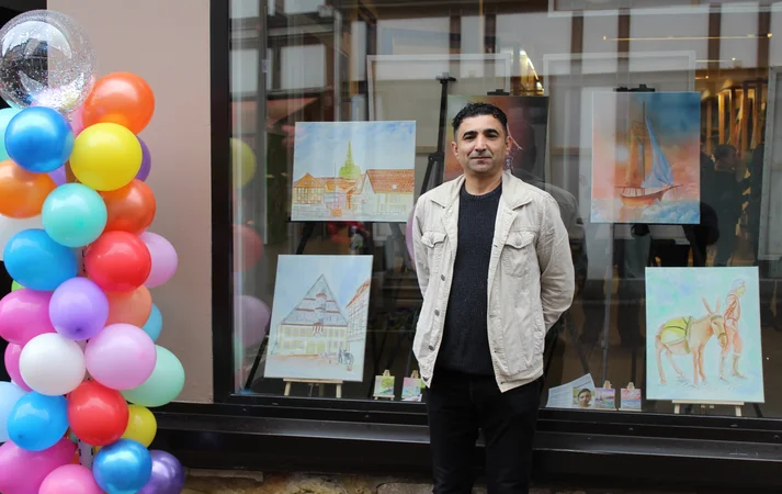 Der Künstler Abdulkarim Alhasan steht vor einem Fenster mit Bildern. Neben ihm sind bunte Luftballons