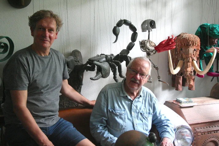 Der Spielemacher Reinhold Wittig mit seinem erwachsenem Sohn Matthias in seinem Zuhause. Hinter Ihnen sind verschidene Marionetten Tiere (ein Skorpion, ein Mammut, etc.) aufgehangen.