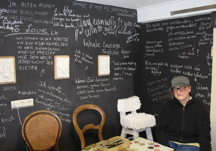 Die Künstlerin Kerstin Schulz sitzt in einem Raum mit Tafelwänden, auf denen mit Kreide von verschiedenen Personen kurue Sätze geschrieben wurden.