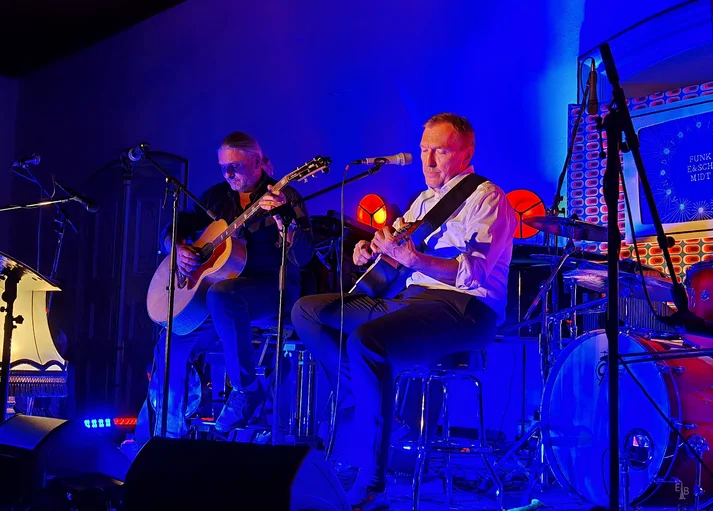 Christian Schmidt und Ronald Gäßlein sitzen nebeneinander auf einer Bühne und spielen beide auf einer Gitarre.