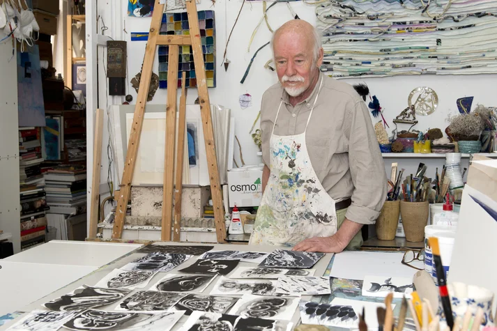 Georg Hoppenstedt steht in seinem Atelier, er trägt eine Malerschürze und blickt auf mehrere Zeichnungen die vor ihm auf dem Tisch liegen.