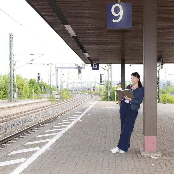 Luise Rist steht auf einem Bahnsteig mit der Nummer 9 und lehnt an einer Säule, sie liest ein Buch.