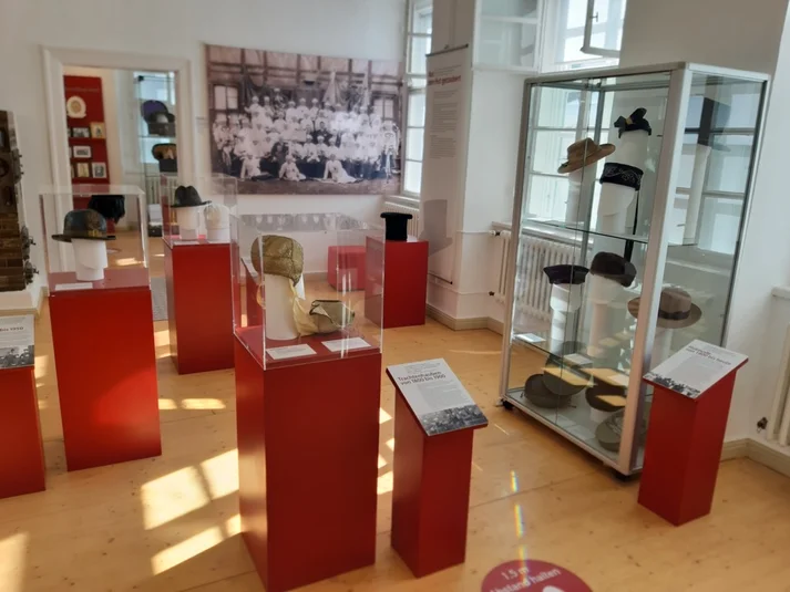 Blick in die Ausstellung Kopfsache im Brotmuseum: Ein heller Raum mit Holzfußboden, darin rote Podeste und Vitrinen mit Hüten