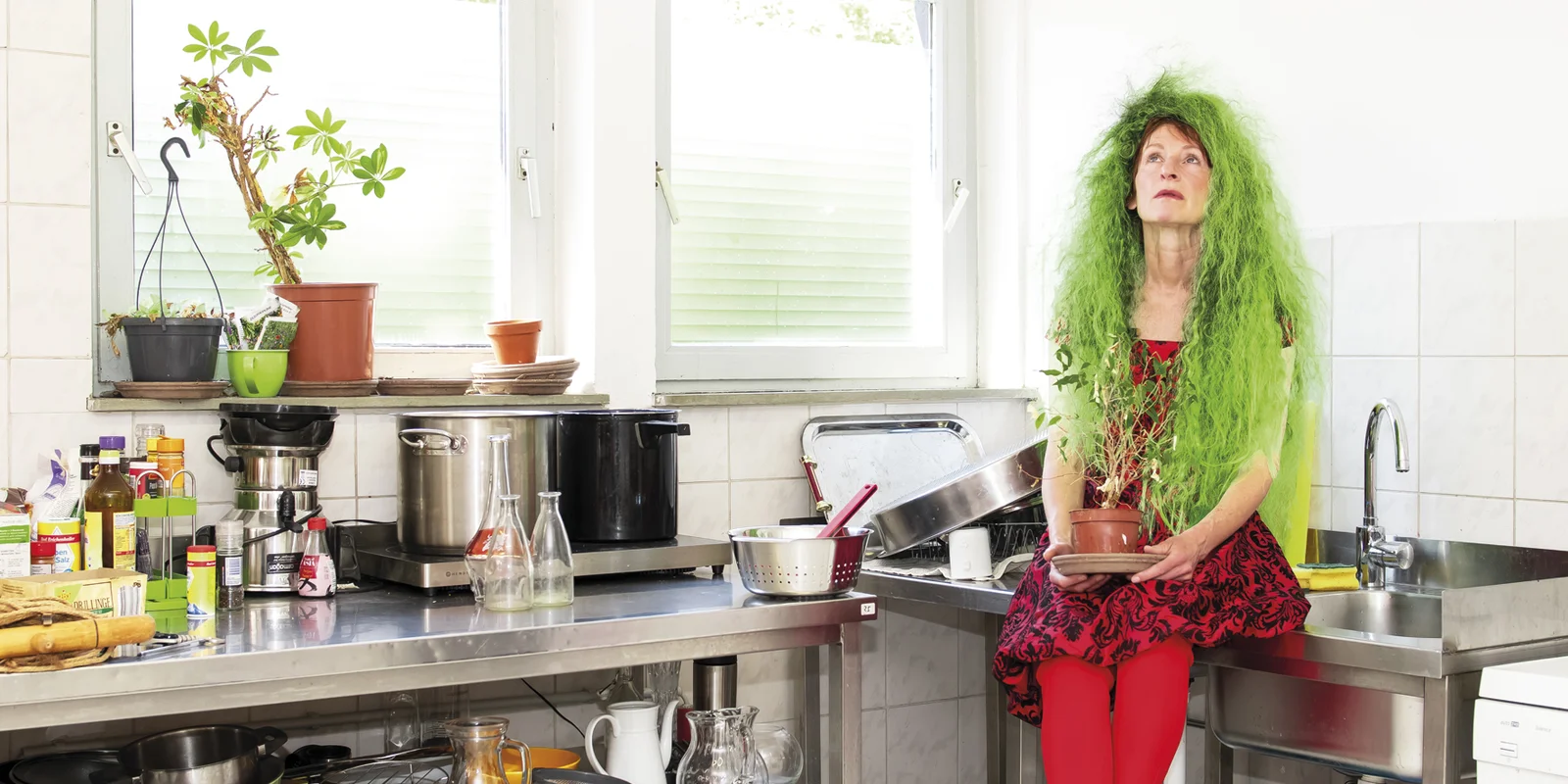 Franziska Aeschlimann sitzt auf einer Edelstahlspüle in einer Küche neben einem vollgestellten Etagenwagen. In der Hand hält sie eine Zimmerpflanze auf dem Kopf hat sie eine Perücke mit langen wilden grünen Haaren.