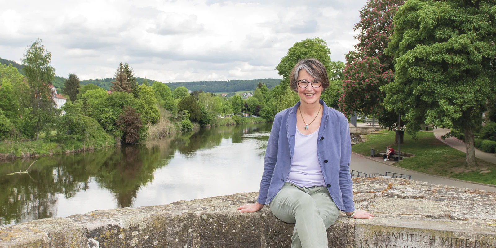 Anja Fehrensen sitzt lächelnd auf einer Steinmauer, im Hintergrund sieht man einen Flusslauf mit grünen Bäumen.