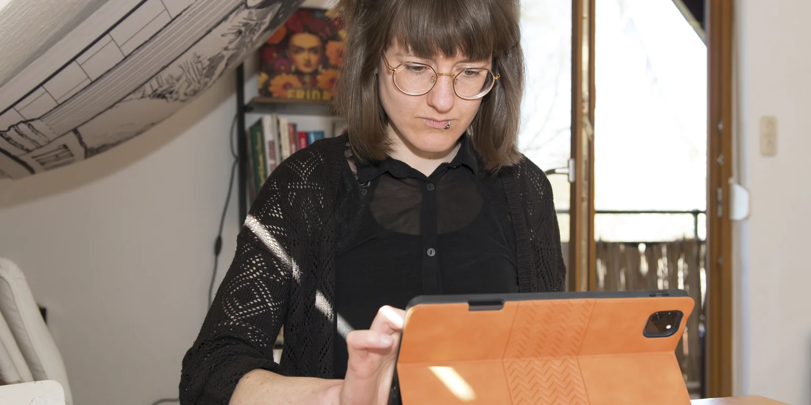 Laura Finke sitzt an einem Tisch auf dem viele Illustrationen liegen und arbeitet an einem Tablet.