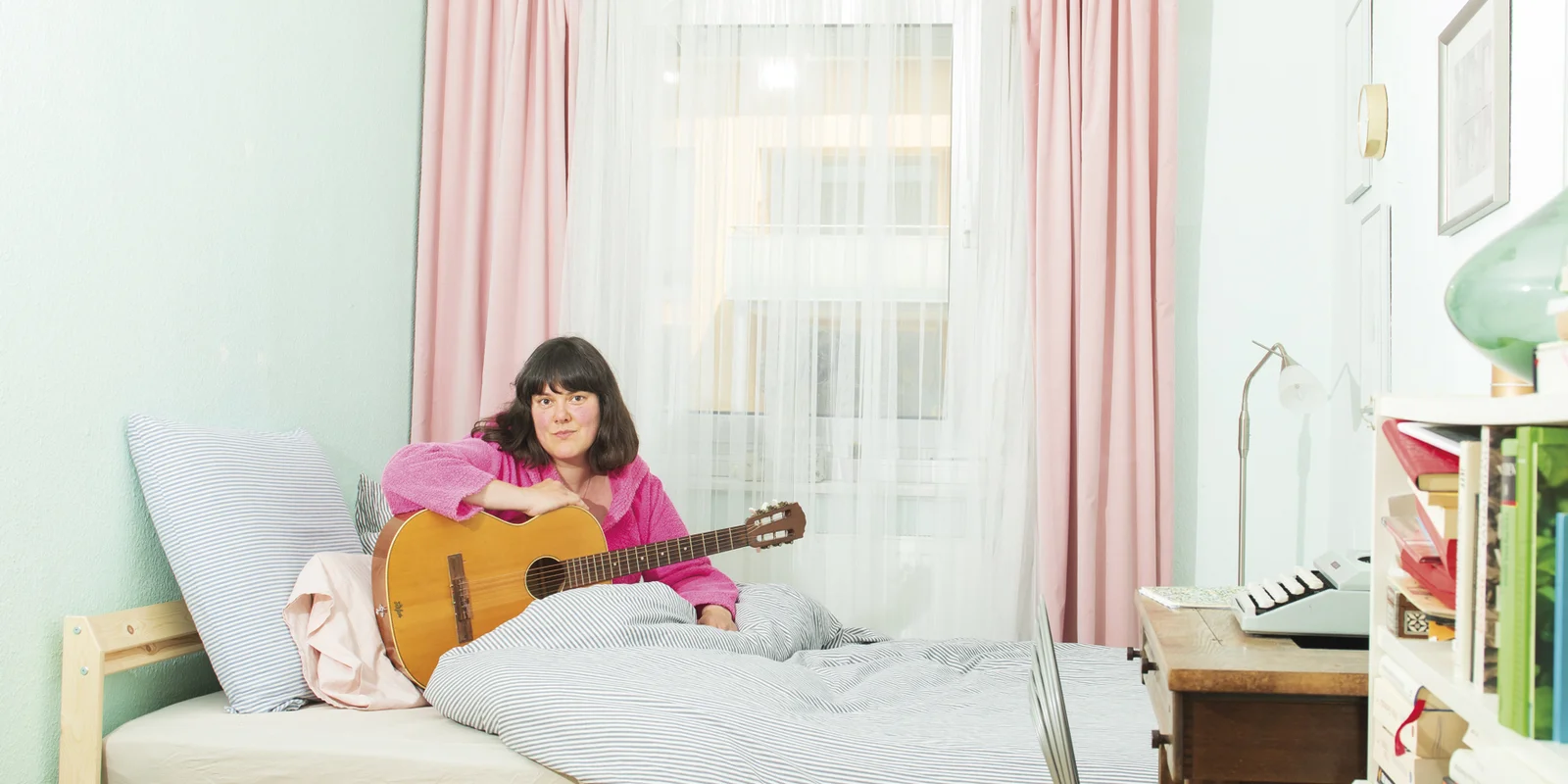 Frau Pauli sitzt im rosa Bademantel auf einem Bett, eine Gitarre vor sich, auf die sie ihren rechten Arm abgelegt hat.