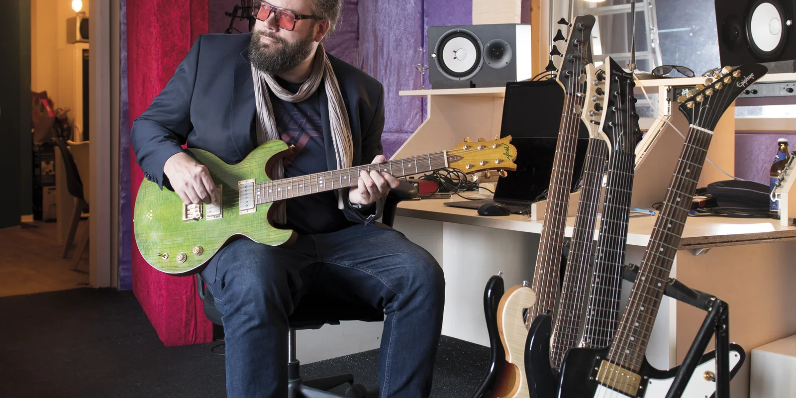 Tobias Langer sitzt mit einer grünen E-Gitarre in der Hand auf einem Barhocker, im Vordergrund sind weitere E-Gitarren auf einem Ständer aufgestellt.