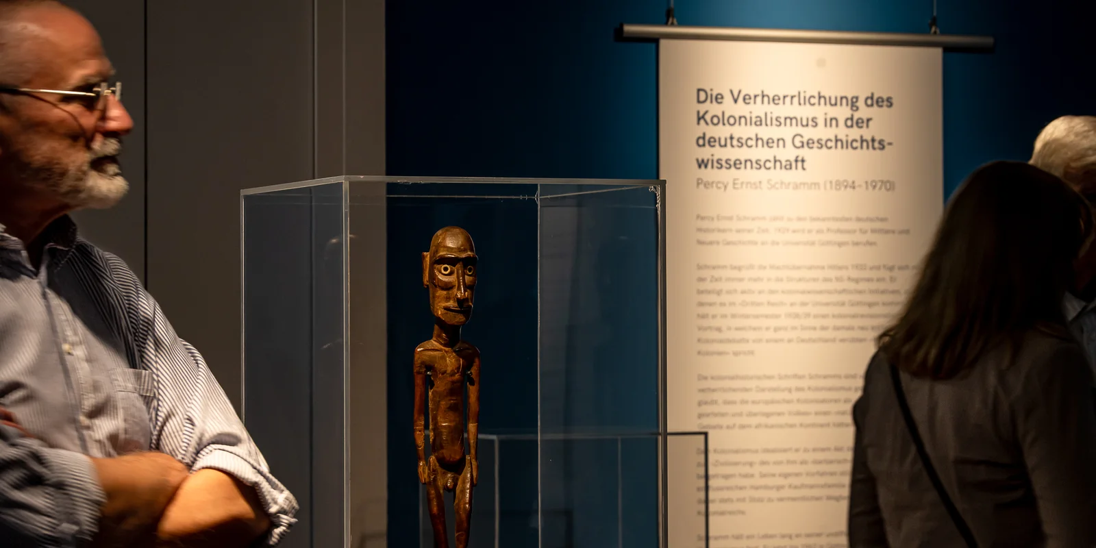 Foto aus der Ausstellung: Göttingen kolonial. Im Vordergrund eine moai tangata (= Holzfigur von Rapa Nui/Osterinseln), im Hintergrund eine Textfahne mit der Überschrift Die Verherrlichung des Kolonialismus in der deutschen Geschichtswissenschaft