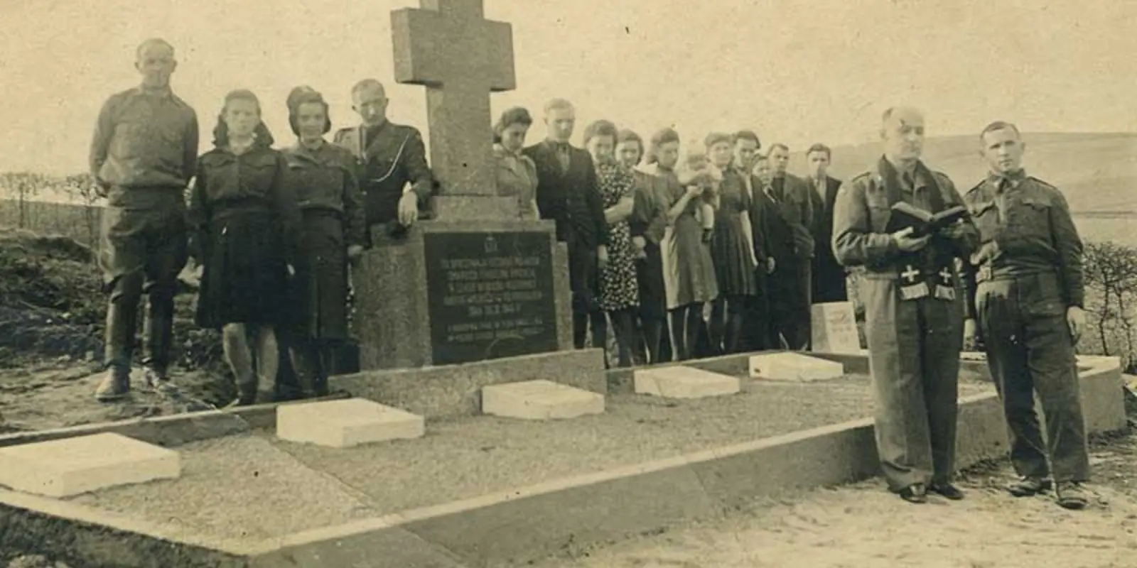 Eine Gruppe von Menschen steht auf dem Schwarz-Weiß Bild neben einem Kreuz auf einem Friedhof hinter einer Grabanlage. Vorne steht ein Priester mit einem Buch.