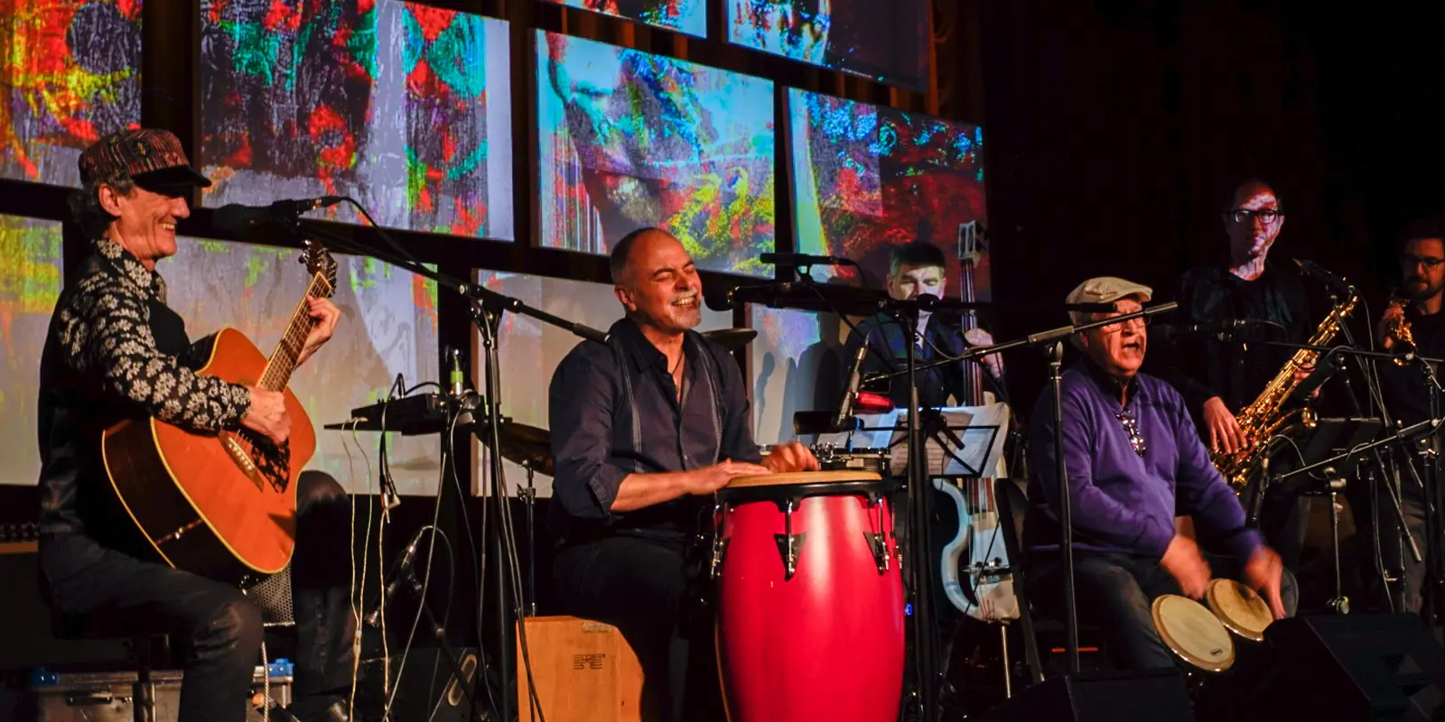 Auf dem Bild zu sehen sind mehrere Musiker*innen mit Gitarre, Schlagzeug und anderen Instrumenten. Im Hintergrund ist eine bunte Videoinstallation zu sehen