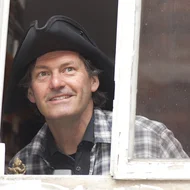 Christoph Buchfink blickt aus einem Fenster mit altem weißem Holzrahmen, er trägt einen dreieckigen Hut.