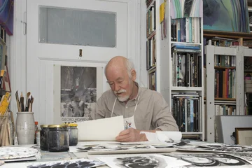 Georg Hoppenstedt sitzt an einem Tisch in seinem Atelier und betrachtet mehrere Bögen mit Zeichnungen.