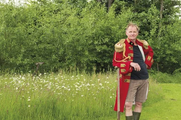 Nik J. Lucht steht auf einen Spaten gestützt in einer roten Uniform und Gummistifeln auf einer teilweise gemähten Wiese.