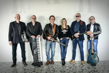 Die Band Rocktail mit 6 Mitgliedern, davon 5 Männer und eine Frau stehen mit ihren Instrumenten in Reihe. Sie tragen alle Jeans und schwarze Jacken.