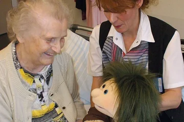 Eine jüngere Frau sitzt neben einer Seniorin und beide schauen auf eine Puppe