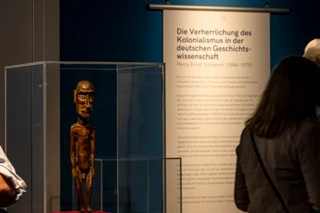 Foto aus der Ausstellung: Göttingen kolonial. Im Vordergrund eine moai tangata (= Holzfigur von Rapa Nui/Osterinseln), im Hintergrund eine Textfahne mit der Überschrift Die Verherrlichung des Kolonialismus in der deutschen Geschichtswissenschaft