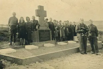 Eine Gruppe von Menschen steht auf dem Schwarz-Weiß Bild neben einem Kreuz auf einem Friedhof hinter einer Grabanlage. Vorne steht ein Priester mit einem Buch.