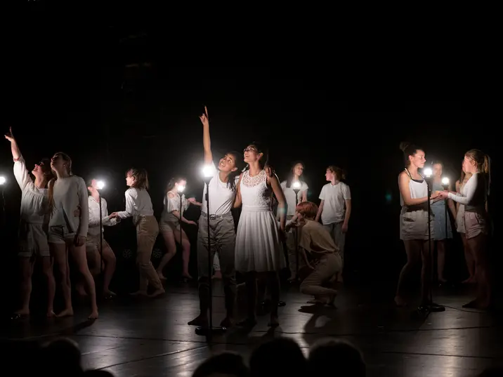 Auf einer abgedunkelten Bühne leuchten kleine weiße Lampen auf Ständern. Schülerinnen des Otto Hahn Gymnasiums sind in Zweierpärchen mit weißen T-Shirt und beigen Hosen oder weisen Kleidern zu sehen. Sie gucken sich gegenseitig in die Augen und halten sich an den Händen. zwei Schülerinnen zeigen mit dem Finger nach oben