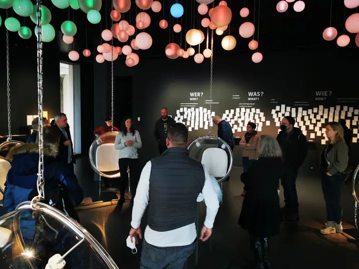 In einem schwarzen Raum hängen Lampen wie grüne, rosa und blaue Luftballons von der Decke. Daneben hängen an Ketten runde Plastikstühle. An der Wand hängen weiße Zettel unter den Überschriften "WER?", "WAS?" und "WIE?". Mehrere Menschen stehen im Kreis und hören einer Frau mit blauem Pullover bei einem Vortrag zu