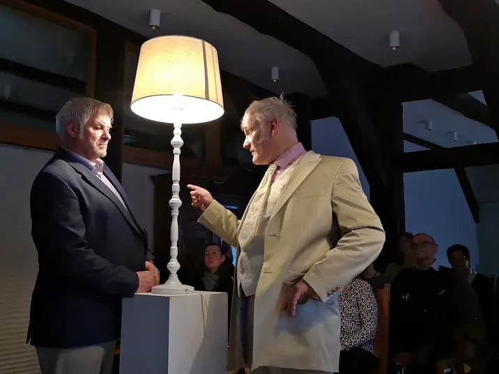 Zwei Männer stehen sich in einem dunkel blauen und beigen Anzug gegenüber. Der rechte Mann zeigt auf den linken Mann. Zwischen Ihnen leuchtet eine Stehlampe.