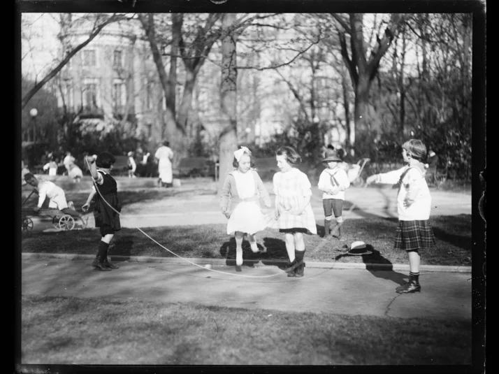 Schwarz-weiß Aufnahme von spielenden Kindern in einem Park im Jahr 1919. Im Vordergrund schwingen zwei Mädchen ein Seil, über das zwei weitere Mädchen springen.