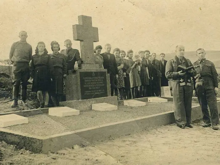 Eine Gruppe von Menschen steht auf dem Schwarz-Weiß Bild neben einem Kreuz auf einem Friedhof hinter einer Grabanlage. Vorne steht ein Priester mit einem Buch.